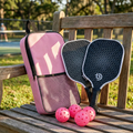 Pink pickleball case with two black pickleball paddles and pink balls on a wooden bench outdoors.
