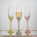 Three ornate champagne flutes with gold, silver, and rose gold bases on a reflective surface.